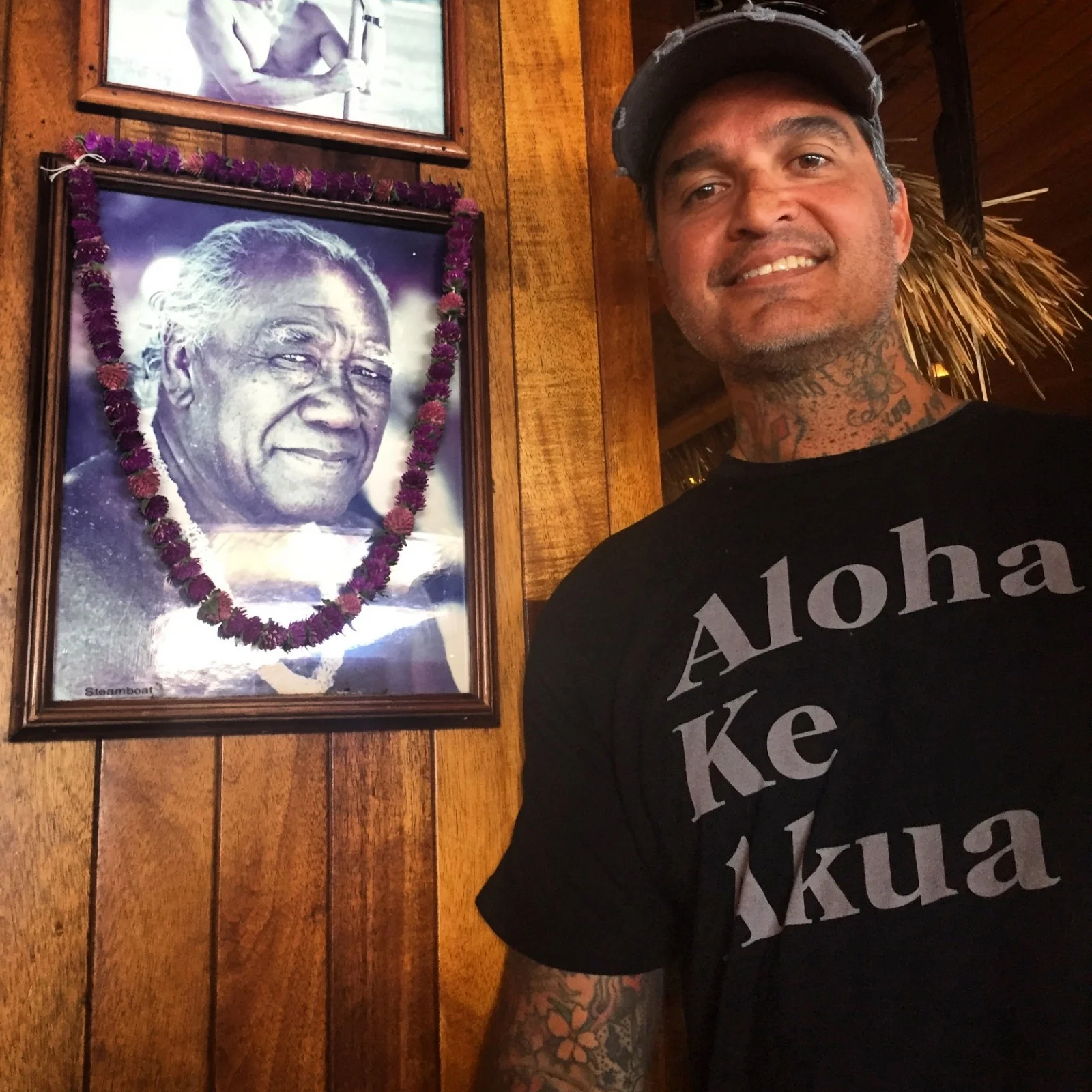 Kevin Mokuahi standing next to his father Sam Steamboat Mokuahi Sr.'s portrait at Duke's Waikiki, wearing an Aloha Ke Akua shirt